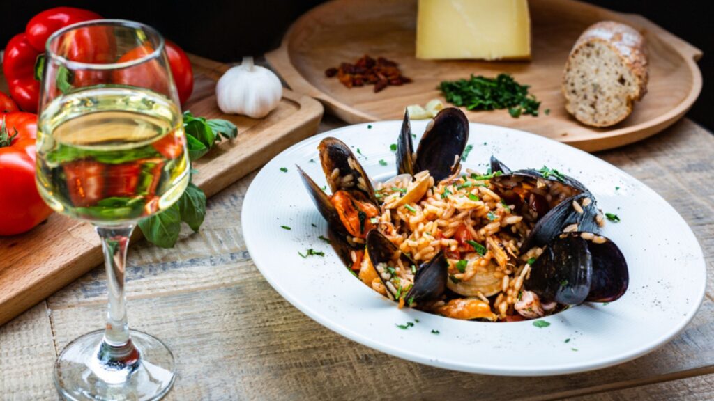 A dish with mussels risotto on a table pairing with a glass of Italian white wine, surrounded by plates with tomatoes, cheese, garlic.
